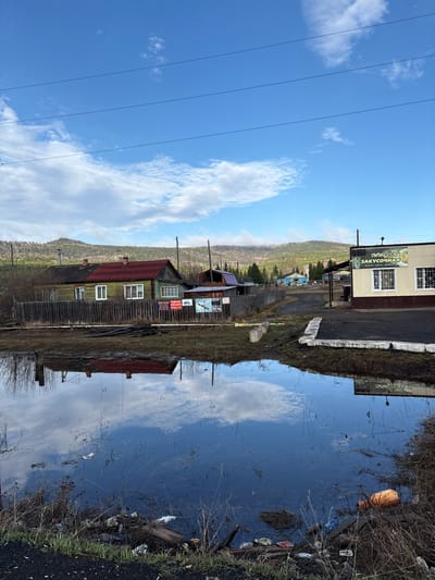 The image shows a rural scene in Sedanovo, Russia. A small body of water reflects the sky and nearby buildings. The buildings appear to be older, wooden structures with various advertisements and signs. A mountain is visible in the background, covered in forest. Power lines stretch across the blue sky with scattered clouds. The water's edge is littered with trash.