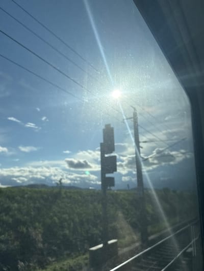 The image, taken from a train window in Loosdorf, Austria, captures a bright, sunny day with scattered clouds. Overhead, four parallel train wires are visible against the blue sky. In the middle ground, two tall poles stand, one with what appears to be a sign or signal box attached. To the left of these poles, a smaller, darker rectangular object is also visible. The landscape consists of a grassy slope leading towards distant, hazy mountains. In the foreground, a section of train tracks runs diagonally from the bottom right towards the left. The sun is bright and positioned high in the sky, causing lens flare and reflections on the window.