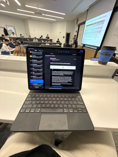 The image depicts an indoor classroom or lecture hall environment, likely located in Wellesley, United States. In the immediate foreground, an iPad attached to a dark-colored Magic Keyboard with a trackpad rests on a light-colored desk, partially obscuring the view of the occupant's light-colored pants and dark top. The iPad screen displays an email application in dark mode, showing an email from "Epic Games" titled "Update to our Terms of Service & Privacy Policy."

Beyond the iPad, approximately 15-20 young adult individuals, presumably students, are seated in tiered rows of light-colored desks, most of whom are observed using personal electronic devices such as laptops or tablets. On the right side of the room, at the front, a male instructor in a blue shirt stands near a lectern or teaching station, facing the class. Behind him, a large projection screen displays a slide containing tabular data and text, partially legible as "competition results in the lowest value." To the right of the screen, a whiteboard shows some handwritten equations. A light blue Caffe Nero coffee cup is positioned on a desk surface to the right of the foreground iPad. The room is illuminated by overhead panel lights, and windows on the far left wall reveal exterior foliage. A red fire alarm pull station is visible on the left wall.