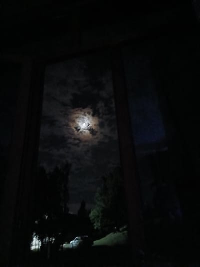 The image shows a view through a dark window frame of a full moon peeking through clouds at night. The moon is bright and prominent in the center of the frame, partially obscured by wispy, illuminated clouds. To the right of the moon, dark silhouettes of trees are visible, with faint lights suggesting some distant structures or activity. The overall scene is dark, suggesting nighttime, and the cloudy sky implies potentially overcast or changeable weather. The view appears to be from an interior space looking outwards, possibly a residential window. There are no visible people or text in the image. The location context provided is Auk, Kyrgyzstan.