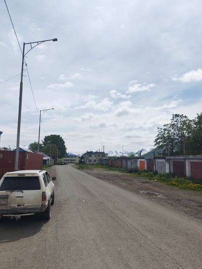 The image captures a street scene in Termal'nyy, Russia, under a cloudy sky. On the left side of the asphalt road, a white SUV is parked, facing away from the camera. Further down the road, several industrial containers are visible. Opposite the containers, a row of garages with various colored doors lines the right side of the road, with green trees and snow-capped mountains in the background. Another street light is positioned on the left side of the road, approximately halfway down. The road surface appears worn, with some puddles and debris present. The overall environment suggests a somewhat utilitarian and undeveloped area.