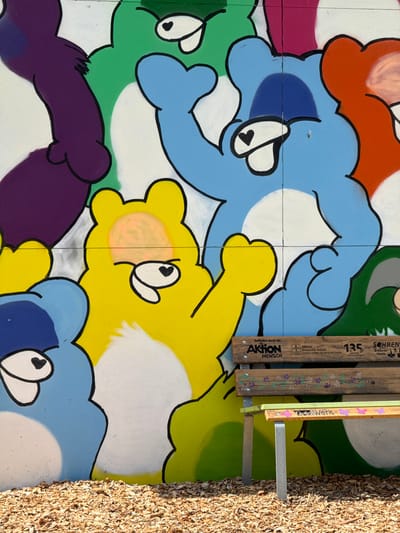 The image depicts a brightly colored mural of cartoon bears on a white wall. The bears are in various shades of purple, green, blue, yellow, and orange. In front of the mural is a wooden bench with metal legs, positioned on a bed of wood chips. The bench has text and logos painted on it, including "AKTION MENSCH" and "135 JAHRE SOHREN". The mural and bench are located outdoors, with the sunlight casting shadows. The overall scene suggests a public art installation or a community space.