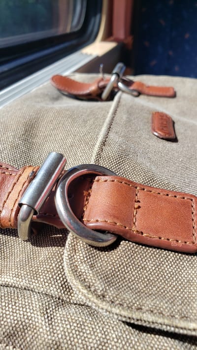The image shows a close-up view of a brown leather strap with a metal buckle attached to a textured fabric bag. The strap is stitched and worn, suggesting it is part of a piece of luggage or a bag. In the background, a window with a blue and brown interior suggests the setting is inside a vehicle, possibly a train, given the perspective and the style of the window frame and seat fabric. The lighting indicates it is daytime, with sunlight casting shadows on the bag. The image does not contain any information that would allow for identification of the city of Hrnčiarovce nad Parnou, Slovakia.