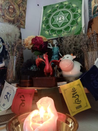 A close-up, eye-level shot focuses on a candle burning in a brass bowl, set on a shelf. Behind the candle, a collection of spiritual and decorative items are arranged. To the left, a white, fluffy seed head sits next to a glass vase filled with dried grass. A red dragon figurine and a black, furry object with eyes are visible further back, partially obscured by a dark brown jar. A statue of a person in a blue robe stands beside a ceramic cow figurine with black spots. In the background, a green mandala-like image is displayed on the wall, alongside a framed picture and a woven tapestry. Small prayer flags, white, red, and yellow, are strung across the foreground, with visible Tibetan script and the word "HUM" on the yellow flag. The lighting is dim and warm, primarily from the candle, creating a mystical and serene atmosphere. The exact location in the city of Пожарское сельское поселение, Ukraine, is not discernible from the image alone.