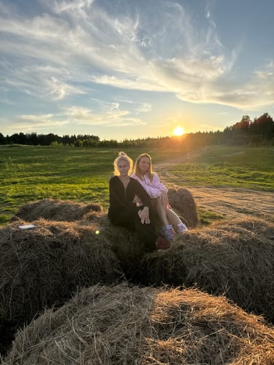 This landscape image captures two women seated on hay bales in a rural field during sunset near Стеклянный, Russia. The sun, a bright golden orb, is low on the horizon to the right, casting long shadows and a warm, golden light across the scene. The sky above transitions from a pale blue to soft orange and yellow hues, streaked with wispy clouds.

In the foreground, two individuals are positioned on large, round hay bales. The woman in front wears a dark long-sleeved top and pants, looking towards the camera with a gentle expression. Behind her, a younger woman or girl in a light pink jacket and light-colored pants with sneakers is also looking at the camera, smiling. They appear to be posing for the picture in a relaxed manner.

The setting is an expansive green field, possibly recently harvested, with additional hay bales visible around the subjects. A dense line of trees or a forest borders the field in the distance, appearing as a dark silhouette against the bright sky. The overall impression is one of peacefulness and natural beauty at dusk. No legible text is visible within the image.