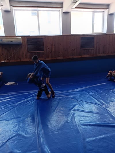 The image depicts two young males engaged in a wrestling match on a blue mat in what appears to be an indoor sports facility. Both individuals are wearing blue uniforms, indicative of a martial art or combat sport. The environment features wooden paneled walls and large windows, suggesting a training hall or gymnasium. The mat has a glossy, reflective surface, and the lighting suggests an overhead source. There are hints of other individuals in the background, also possibly participants, but they are out of focus. The overall scene captures a moment of physical exertion and training. The location is identified as Ladyzhyn, Ukraine, though specific landmarks are not visible within the frame.