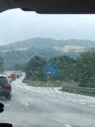 The image captures a view from inside a car on a highway in Hvězdonice, Czechia, during a rainy day. The foreground is dominated by streaks of rainwater on the windshield, distorting the view of the road ahead. Several vehicles are visible on the highway, including a black car in the left lane and red trucks further up the road, suggesting a traffic slowdown or congestion.
The setting is a rural highway with lush green trees lining the sides of the road and rolling hills in the background. The sky appears overcast, contributing to the gloomy weather. The time of day is difficult to pinpoint precisely due to the weather conditions but appears to be daytime, likely mid-afternoon or late morning.
A blue road sign is partially visible on the right side of the highway. While the text is blurred by the rain and the image resolution, it appears to be a directional sign with some lettering. Below the sign, there are warning signs indicating a curve in the road. The overall mood conveyed is one of a typical, somewhat dreary travel day, impacted by inclement weather.