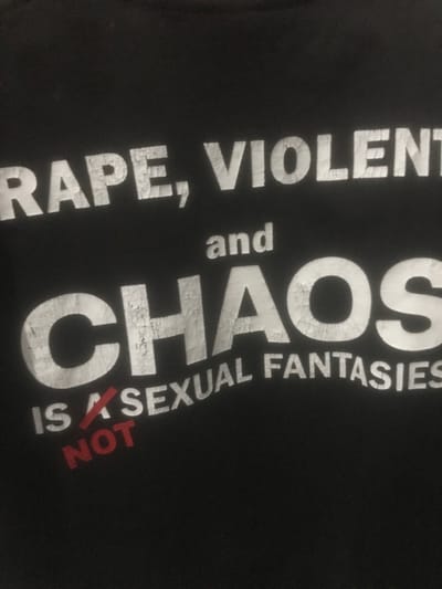The image displays a close-up of a black fabric, likely a t-shirt, printed with white and red text. The text is slightly angled upwards from left to right. The white text, appearing somewhat distressed, reads: "RAPE, VIOLENT" (where "VIOLENT" is likely part of "VIOLENCE" and is partially cropped on the right), followed by "and" on the next line, then "CHAOS" in a larger, bolder font. Below "CHAOS," the white text continues: "IS A SEXUAL FANTASIES." The phrase "IS A" is deliberately crossed out with a diagonal red line, and the word "NOT" is printed in red directly beneath it. No individuals, actions, interactions, or discernible environmental features are visible. The specific location in Limbangan Sari, Indonesia, cannot be determined from this image.