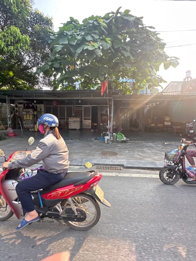 The image depicts a bright, sunny street scene in Vị Xuyên Commune, Vietnam, likely during the golden hour, characterized by strong sunlight and long shadows.
In the immediate foreground, a person is seated on a red Honda RSX scooter, positioned on the right side of the frame. The rider wears a grey long-sleeved jacket, dark blue trousers, and blue sandals. A blue helmet, featuring the letters "GP", is worn on their head, and a pink face mask covers their lower face. The scooter's license plate is partially visible, showing "89-F1 006.27".
Further up the street on the right, another person is seen on a stationary electric bicycle with a front basket, facing away from the viewer.
The street itself is paved with concrete or asphalt, displaying a manhole cover and faint road markings. On the left side of the street, a row of buildings, presumably shops or residences, features open fronts. Several signs are visible on these establishments; one prominently displays "HAU", and another reads "DO DONG HO" and "DO NHIEU". A partial sign reads "CUA HANG". A red flag is positioned near one of these buildings, and a green bucket and a white bucket are on the sidewalk.
Lush green trees line the street, and utility poles with overhead wires are present. The scene captures a typical, vibrant street environment.