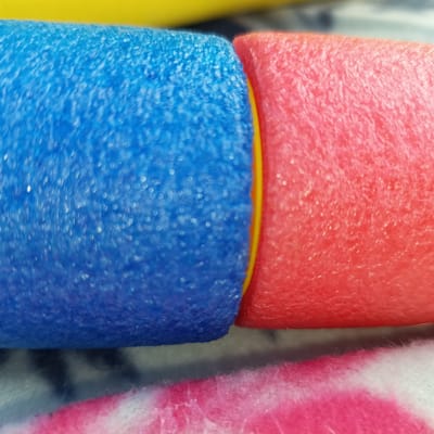 The media file shows a close-up, macro shot of two foam pool noodles, one blue and one red, joined together by a yellow connector. The noodles are textured and appear to be made of foam. The background is blurred, showing a hint of a pink and white patterned fabric or surface below. The lighting suggests an indoor or overcast outdoor setting. There is no visible text, people, or discernible activity. The focus is entirely on the texture and colors of the pool noodles.