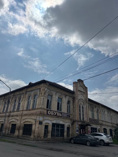 The image captures an exterior view of a weathered, yellow brick building in Nogornyi, Russia, under a partially cloudy sky. The building exhibits architectural details such as ornate window frames and decorative brickwork. A sign above a storefront reads "ОБУВЬ" (Shoes) in large letters, with smaller text below indicating services like "ALL FOR THE FAMILY - FOR ORDER - ALL TYPES OF REPAIR." Several windows on the ground floor are covered with metal grates. Two cars, a dark sedan and a white SUV, are parked on the street in front of the building. Overhead, several power lines are visible against the sky. The environment appears to be an urban street scene.