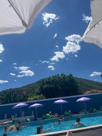 In Varvara, Bulgaria, this image captures a poolside scene under a clear blue sky with scattered white clouds. Two large white umbrellas are visible in the foreground, partially obscuring the sky. A vibrant blue swimming pool occupies the lower half of the frame, with several people swimming or relaxing in and around it. Green beanbag chairs are scattered on the pool deck, some with people seated on them. Two purple umbrellas with "Ice Cream" branding are planted behind the pool. In the background, a lush green, tree-covered mountain rises, with red-tiled roofs of houses peeking through the foliage. The overall atmosphere suggests a sunny day at a resort or a recreational area.