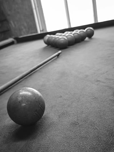 The image captures an indoor scene in Терновский округ, Ukraine, rendered in monochrome. The primary subject is a billiard table covered with textured felt. In the foreground, a single cue ball exhibiting visible wear and markings, appearing to be "III" or "111", is positioned on the table's surface. A billiard cue lies horizontally on the felt, extending from the left mid-frame towards the upper right. Further down the table, a triangular rack containing approximately 15 to 16 additional billiard balls is arranged near the far end. The background features a large window with vertical divisions, admitting intense natural light that results in an overexposed exterior view. A dark, indistinct wall or structural element is visible to the left of the window. No individuals are present in the scene, which depicts a static arrangement of billiard equipment.