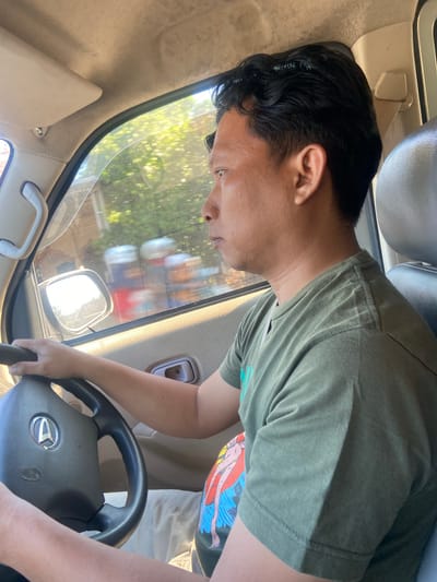 An Indonesian man is driving a vehicle in Ngrame, Indonesia. He is wearing a green t-shirt and has dark hair. The interior of the vehicle is visible, including the steering wheel, dashboard, and side mirror. The scene appears to be during daylight hours, with sunlight filtering through the windows. The man's hands are on the steering wheel as he looks forward. The background outside the window shows blurred foliage and structures, suggesting movement.