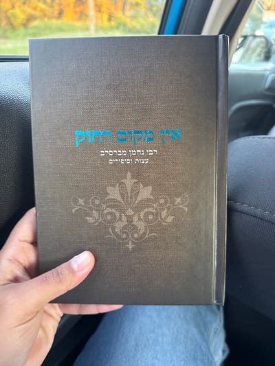 A left hand holds a dark gray, textured hardcover book, titled in bright blue-green Hebrew script: "אין מקום רחוק" (There is no distant place), "רבי נחמן מברסלב" (Rebbe Nachman of Breslov), and "עצות וסיפורים" (Advice and Stories). A faint, darker gray stylized floral design is centered below the text on the book's cover. The book rests on the lap of a person wearing blue denim jeans. The scene is inside a vehicle, indicated by a dark gray fabric car seat on the right and a light blue interior trim piece in the upper left. Through the vehicle's window, out of focus, are autumnal trees with orange and green foliage under daylight conditions. The location is West Windsor, United States.