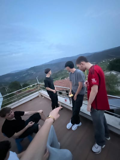 The image shows a group of young men on a rooftop in Celorico de Basto, Portugal. Three are standing near the edge, with one wearing a red shirt, another in a grey shirt holding a cell phone, and the third in black. One is pointing at the group with an arm and a gold watch showing. Two other people are sitting on the floor. The background features mountains and a cloudy sky. Buildings are visible below. A safety railing lines the roof's edge.