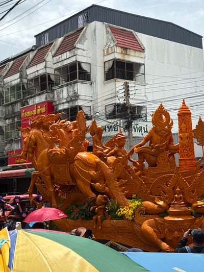 The image depicts a street scene in Prakhon Chai District, Thailand, during what appears to be a parade or festival. A large, intricately carved orange float, likely made of wax, dominates the foreground. The float features mythical creatures, including a multi-headed horse with riders and elaborate decorative elements. Behind the float, a row of multi-story buildings with weathered facades and red tiled roofs line the street. Overhead, power lines crisscross the sky, indicating an urban setting. In the lower portion of the image, the tops of colorful umbrellas and crowds of people are visible, suggesting a public gathering. One person in the lower right corner is taking a photograph of the float with a mobile phone.