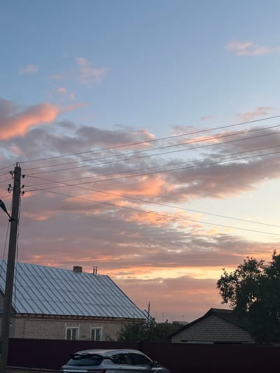 This is a vertical, eye-level, medium shot of a rural sunset scene in the Сароцкі сельскі Савет, Belarus. The sky is filled with pastel-colored clouds, transitioning from soft pinks and oranges near the horizon to pale blue at the top. Power lines stretch horizontally across the frame, supported by a wooden utility pole on the left. In the foreground, the roof of a light-colored house with a metal roof is visible. A dark fence runs along the bottom of the image. A dark-colored vehicle is parked in front of the fence. To the right, the dark silhouette of a tree partially obscures a smaller brick building. The scene is devoid of any human presence.