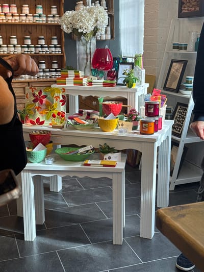 This image presents a forensic view of a retail interior located in Hornsea, United Kingdom. In the central foreground, two white display tables with fluted legs are arranged, with a taller table positioned behind a shorter one. The upper display table exhibits a square decorative glass panel featuring multicolored botanical designs, stacks of brightly colored notebooks, a tall clear glass vase with white artificial hydrangeas, a red glass vase, various patterned ceramic bowls (red, yellow), and multiple jarred candles (pink, orange). The lower table displays additional patterned ceramic bowls (mint green, red), a green leaf-shaped bowl, and small tea light holders.
To the far left, the partial view of an individual's left arm and torso is visible, clad in a dark garment, with a blurred handheld device near their waist. A wooden shelving unit in the left background is fully stocked with numerous small, uniformly sized jars, likely containing paints or craft supplies. In the right background, a white ladder-style shelving unit holds framed artwork (including one labeled "BAUHAUS") and assorted decorative bowls. A second individual's lower right leg and foot, wearing dark trousers and a navy shoe, are visible on the far right. The floor is composed of large dark grey square tiles with lighter grout lines. A window with a sheer white curtain is visible on the upper right wall. The scene is well-lit, casting distinct shadows on the tiled floor.