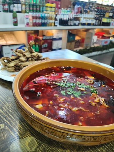A close-up, eye-level shot captures a bowl of spicy Chinese stew, possibly fish or meat, in a rustic ceramic bowl with a decorative rim, resting on a wooden table. The stew is a vibrant red, indicative of chili oil, and contains visible pieces of protein, dark ingredients like mushrooms, and is garnished with fresh green herbs, likely cilantro. To the left of the bowl, a plate holds a pile of golden-brown, crispy fried food, possibly fish or vegetables. In the background, shelves are stocked with an assortment of beverages, including bottles of beer and various soft drinks, suggesting the setting is a restaurant or bar in Jixi, China, a city known for its coal mining and developing tourism. The lighting appears to be artificial, illuminating the food and creating a warm ambiance.