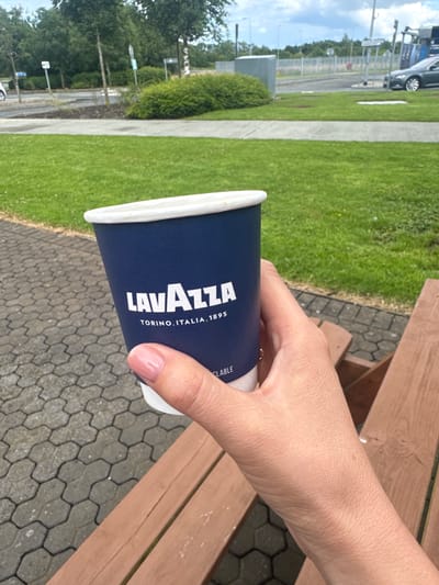 A hand with light skin, a neatly manicured light pink nail, and a thin gold ring on the index finger holds a dark blue disposable coffee cup with a white rim. The cup has "LAVAZZA TORINO, ITALIA, 1895" printed in white text, with the "LAVAZZA" text larger and more prominent. A smaller line of text "CLABLE" is also visible to the right of "LAVAZZA".

The setting appears to be outdoors, with a wooden bench visible to the right of the cup, suggesting a seating area. The background includes a paved area with hexagonal interlocking stones, a patch of green grass, and a grey concrete path. Further in the background, there are trees, streetlights, road signs, and a glimpse of a dark-colored car and what looks like a fenced-off area, possibly a parking lot or a secure facility. The sky is visible with clouds, indicating daytime. The overall mood is casual and relaxed, likely a moment of enjoying a coffee break outdoors. The location context places this scene at Commons Cross, Ireland.
