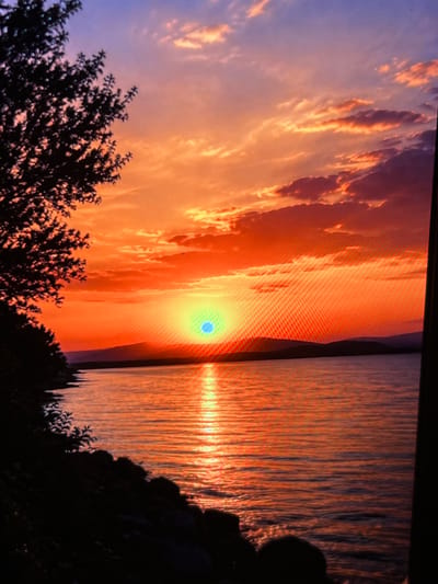 This image depicts a vibrant sunset over a body of water, likely a lake or a wide river. The sky is a dramatic canvas of oranges, reds, and purples, with fluffy clouds catching the warm light. The sun, appearing as a bright, almost turquoise orb, is low on the horizon, casting a shimmering reflection on the water's surface. Silhouetted against the bright sky are dark, undulating hills in the distance.
In the foreground on the left, the dark silhouette of a tree with dense foliage overhangs the shoreline. The shoreline itself is lined with dark rocks and vegetation, also rendered in shadow. The water exhibits gentle ripples, reflecting the intense colors of the sunset.
The image has a visible pixelated quality, suggesting it may be a photograph taken of a screen or a digital image with a low resolution.
There is no visible indication of the city of Montfermeil, France, in this image, nor are there any people or specific actions being depicted. The scene is purely naturalistic, focusing on the atmospheric beauty of a sunset.
