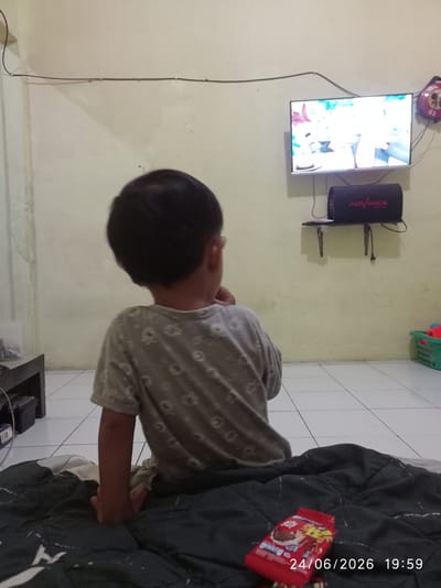 The image shows a young child, seen from behind, engrossed in watching a television. The child is wearing a light-colored t-shirt with a subtle pattern and is seated on a dark comforter. In front of the child, on the comforter, is a red wrapper with visible text and an image of what appears to be a snack. The television is mounted on the wall, displaying a bright image, likely a cartoon or animated program. Below the TV, a black speaker with the word "ADVANCE" in red lettering is visible, mounted on a small shelf. The room appears to be a living area with tiled flooring. A green plastic basket, possibly containing toys, is visible in the background to the right. The overall lighting suggests it is evening, further confirmed by the timestamp "24/06/2026 19:59" superimposed on the image. The scene depicts a common domestic setting, with the child fully absorbed in entertainment. The location context is Sukaraja, Indonesia.