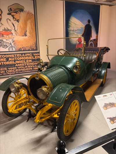 An indoor scene, likely a museum or exhibition in Wassenaar, Netherlands, showcases a meticulously restored vintage dark green automobile with bright yellow spoked wheels and brass accents. The car, identified by a display board as a "1909 GEORGES ROY 12-HP TOURING SPORT TORPEDO", features prominent external headlamps, an engine with horizontal fins, and a spare yellow wheel mounted on its side. Its interior, partly visible, includes dark seating and a wooden steering area.

Two large framed posters adorn the wall behind the car. The poster on the left depicts a man in a hat smoking a pipe, holding a magnifying glass over a small car. Partial Dutch text reads: "TOONSTELLING VAN... AUTOMOBIELEN... AMSTERDAM... 21 DECEMBER 191[X]... De Rijwielen en Automobiel:Indust," indicating an early 20th-century automobile exhibition. The poster above the car shows a figure in a dark coat looking into a beam of light that illuminates a distant structure, possibly a castle. A red object is partially visible in the foreground of this poster.

Adjacent to the car, a white display board provides further details, including the car's engine specifications ("17-LITRE FOUR CYLINDER POWER 12 HP") and notes in both Dutch and English about its configuration, such as "Georges Roy als tweezitter" and "Georges Roy as a two seater." The floor is made of light-colored tiles, and the overall lighting suggests an artificial, controlled environment typical of a museum. The scene captures a moment of historical automotive display.