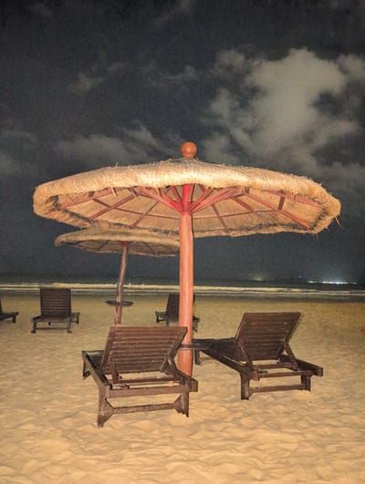 The image shows a sandy beach at night with two large thatched umbrellas and several wooden lounge chairs. The sky is dark and cloudy, with some distant lights visible along the horizon, likely indicating a town or city. The ocean waves are visible in the background. The lounge chairs are made of dark wood and are arranged on the sand. One umbrella is in the foreground, and another is partially visible behind it. The umbrellas have red poles and thatched tops. The scene appears to be peaceful and serene, with no people present. The location is identified as Tianya District, China.