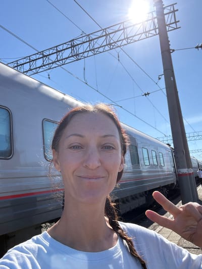 A forensic description of the scene depicts a single adult female in the foreground, centered and looking directly at the viewer with a slight smile. She has light skin, dark hair styled in a braid over her right shoulder, and is wearing a light-colored, long-sleeved garment. Her left hand is raised, displaying a "peace" sign (V-sign) with her index and middle fingers extended.
The background features a silver-grey passenger train with multiple rectangular windows, running horizontally across the middle ground. A red stripe is visible along the lower portion of the train's body. Overhead, a clear blue sky dominates, with a bright sunburst emanating from the upper right, casting a strong light. An extensive railway electrification system, consisting of dark metal trusses, wires, and a tall vertical pole, is visible against the sky and behind the train. In the far distance to the right, beyond the train and the pole, faint figures of other individuals are discernible on what appears to be a railway platform. The location is a train station or railway stop in Kropachyovo, Russia.