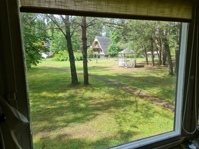 This is a daytime, eye-level, exterior shot taken from within a building, looking out of a window onto a grassy yard in Kirneilė, Lithuania. The yard is dominated by a well-maintained lawn, with several trees scattered throughout. In the background, a distinctive A-frame house with a dark-pitched roof is visible. To the right of the house, a white wooden gazebo with a peaked roof stands in the yard. In the middle ground, a stone pathway winds through the grass. A children's swing set with a slide is also present, near the base of two prominent trees in the foreground. The window frame and a portion of a light-colored blind are visible in the foreground, obscuring the left side of the view. The overall impression is of a serene, rural setting.