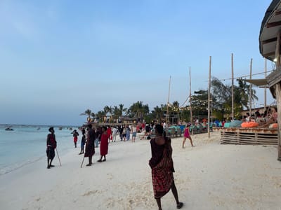 The image depicts an active beach scene in Nungwi, Tanzania, observed during dusk or early evening hours. The sky above is a clear, light blue hue. The foreground consists of white sand beach transitioning into shallow, clear turquoise ocean water. Several small boats are anchored further out in the water on the left, with one jet ski visible near the shore.
In the immediate foreground, a dark-skinned individual wearing a red and black patterned wrap garment is positioned with their back to the camera. To the left, near the water's edge, three other dark-skinned individuals, similarly dressed in red and black patterned wraps and each holding a wooden stick, are standing in various orientations.
The mid-ground of the beach is densely populated with a diverse group of people, including adults and children, walking, standing, and engaging in social interactions. Many individuals are dressed in casual beach attire. On the right side of the frame, a raised wooden platform functions as a lounge area, furnished with multiple orange and white bean bag seats occupied by patrons. Behind this, a series of beachfront establishments constructed primarily from wood are visible, illuminated by strings of overhead lights. These establishments feature numerous tables, some covered with blue and red cloths, with chairs and seated individuals. Palm trees line the perimeter of these structures. A wooden pier extends into the ocean on the far left of the frame.