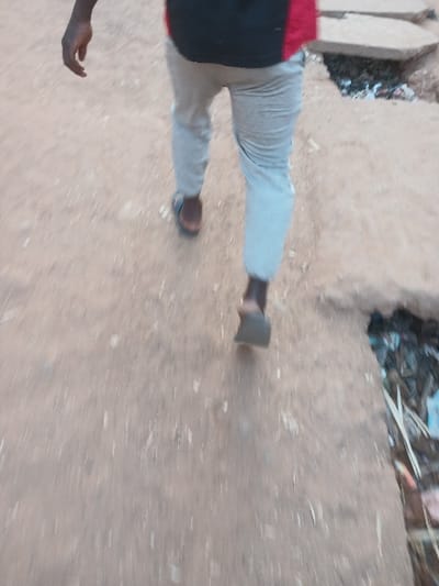 The image, taken in Dogon Nama, Nigeria, depicts a person walking on an unpaved, light brown dirt path or ground. The photograph appears to be rotated 90 degrees clockwise, placing the walking individual diagonally across the frame.
The main subject is a person, partially visible from the torso down. They are wearing light-colored trousers, likely grey or light blue, and a dark top with a red section visible on the upper left side of their body. On their feet, they wear simple, dark-colored sandals. The person is in motion, captured mid-stride, with one foot on the ground and the other lifted, indicating they are walking. A dark-skinned hand, with fingers slightly curled, is visible in the lower left portion of the image.
The setting is an outdoor area characterized by loose, sandy or dusty ground. Small pebbles or debris are scattered across the path. In the background, particularly towards the upper right (if the image were oriented vertically), there is a cluttered area containing dark refuse, possibly garbage or organic debris, piled against what seems to be a low wall or raised bank.
The activity taking place is simply walking. The lighting suggests it is daytime, with soft, diffused light, as there are no distinct shadows. No specific weather conditions are discernible, but the dry, dusty ground implies fair weather. There is no visible text in the image, and no specific emotions can be discerned from the partial view of the person. The overall scene presents an everyday moment in what appears to be a natural or semi-urban outdoor environment.