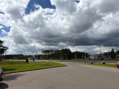 This image depicts a roadside scene in Byerazino, Belarus, under a dramatic sky filled with dark, brooding clouds interspersed with patches of blue. The time of day appears to be afternoon, given the light.
The foreground is dominated by a paved road or parking area. To the left, the rear quarter of a silver car is visible. Beyond the pavement, a strip of vibrant green grass slopes upwards, dotted with small yellow wildflowers and a few large rocks. A young, dark green evergreen tree stands alone on the grass.
In the middle ground, a road runs horizontally, bordered by a line of tall, dark green pine trees that form a dense forest. Beyond this forest, several buildings are visible, including what appears to be a gas station with prominent signage, and a larger, multi-story building with a light-colored facade. Vehicles, including cars and trucks, are present at the gas station and on the road.
The sky is a significant element, with heavy, grey clouds covering most of the upper frame, suggesting an impending or recent rain shower. The overall mood is somewhat somber due to the weather, yet the lush green of the grass and trees provides a sense of natural beauty.
A blue and white sign with Cyrillic lettering, likely a gas station logo and information, is clearly visible on the right side of the middle ground. There are also traffic signs visible on the road, including a circular red sign with a white horizontal bar, indicating "no entry" or "do not enter." The presence of the gas station and the road layout suggest a commercial or travel hub.