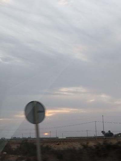A muted landscape features a signpost with a horizontal line at left. Beyond this, a harvested field stretches to the horizon under a gray, overcast sky. Distant power lines traverse the scene. A pale sun, low in the sky, peeks through a gap in the clouds, creating a thin bright strip. Dark vegetation dots the horizon. The viewpoint is low, possibly from a vehicle.