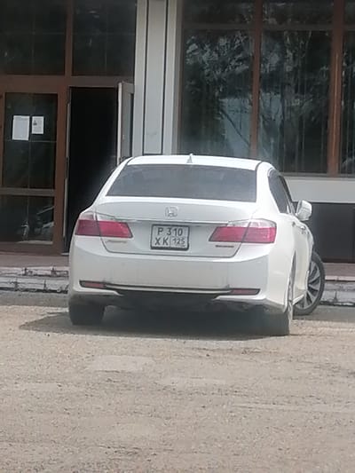 A white Honda Accord is parked on a gravel lot in front of a building with large windows and a dark doorway. The car's license plate, which reads "P 310 XK 125," is visible. The building appears to be an office or commercial establishment. The scene is set in Lesozavodsk, Russia.