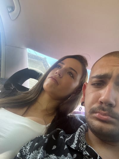 A close-up, tilted selfie shows two people inside a car. The man is positioned at the top of the frame, partially cut off, with dark hair, a beard, and wearing a black patterned shirt. A small, dark earring is visible in his left ear. The woman is below him, wearing a white top and a silver hoop earring in her left ear. The car's interior is light-colored, and part of the window is visible behind the woman. Outside the window, glimpses of a building can be seen. The specific location in Zlatna Panega, Bulgaria, is not discernible from the image.