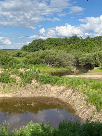 A yellow boat with two people on board navigates a narrow river surrounded by lush green foliage and trees under a bright blue sky with fluffy white clouds. The riverbank in the foreground is sandy with dry vegetation. The background features a dense forest on a rolling hillside, indicating a natural, possibly rural or park-like setting. The location is identified as Маяк, Russia.
