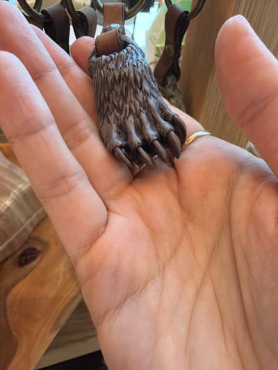 A person holds a dark brown bear claw keychain in their left palm. The hand features a gold ring. The background includes wood surfaces and other parts of the keychain. Location: Cannot be determined from the image.
