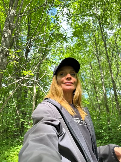 A medium shot captures a woman from the chest up, facing the camera with a slight smile. She wears a black baseball cap, a grey zip-up jacket over a black t-shirt, and her long blonde hair falls around her shoulders. The background is a dense forest with tall trees and vibrant green foliage, suggesting an outdoor, natural setting. The lighting indicates it is daytime, with sunlight filtering through the leaves, creating a dappled effect. The woman appears to be standing or walking in the forest. The location is specified as Interkompleks, Russia.