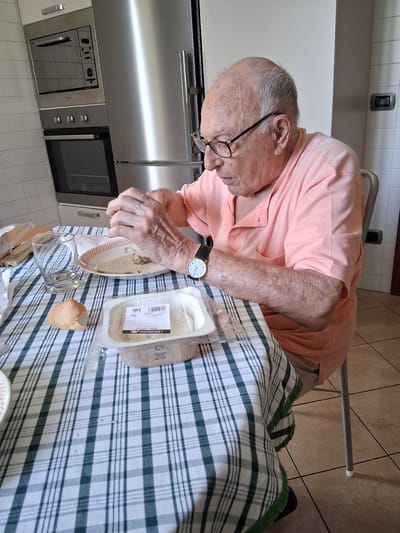 The image shows an elderly man with grey hair and glasses sitting at a table covered with a green and white checkered tablecloth. He is wearing a light pink polo shirt and a wristwatch. The man is holding a fork and appears to be eating from a small plate with some food on it. In front of him, there is a plastic container of what looks like cheese, a small bread roll, and a glass.

Behind the man, part of a kitchen is visible, including a stainless steel refrigerator and built-in ovens. The floor is tiled, and the overall impression is that of a domestic setting, likely a home kitchen. The lighting suggests it is daytime. The man's focus is on his meal, and his expression is neutral. There is some text visible on the plastic packaging, but it is not legible enough to read. The context provided indicates the location is Padua, Italy.