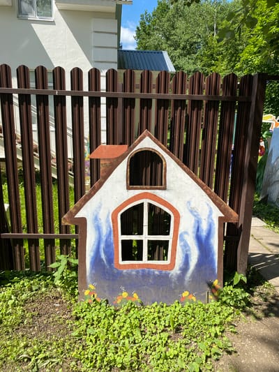 A colorful, cutout representation of a house stands in front of a dark brown, corrugated metal fence. The cutout house is painted white with blue flames extending upwards from the bottom, and orange trim around its windows and roofline. Small yellow and orange flowers are painted at the base of the cutout. The fence is made of vertical corrugated metal panels. Behind the fence, a white building with decorative molding and a window is visible. Trees and greenery are present in the background, suggesting an outdoor setting. The scene appears to be in a park or garden area.