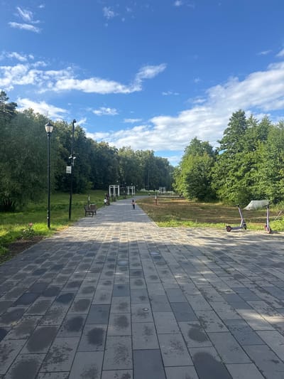 The image depicts a paved park walkway in Sarov, Russia, under a bright blue sky with scattered white clouds. The walkway is made of rectangular grey pavers, some of which appear wet, suggesting recent rain or watering. To the left of the walkway is a grassy area with trees, a lamppost, and a bench. Further down the walkway, there is a child on a scooter and another child standing near a bench with an adult. To the right of the walkway, more trees and a grassy area are visible, with two purple electric scooters parked. In the background, there are white structures that resemble gazebos or pergolas, and a dense line of trees forming the park's boundary. The overall scene is one of a well-maintained urban park on a pleasant day.
