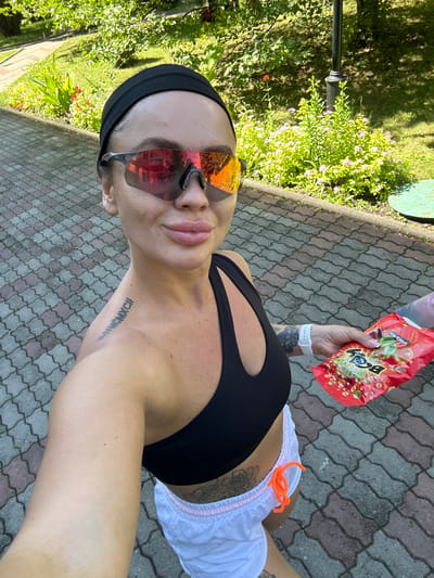 A woman is taking a selfie outdoors. She is wearing a black headband, mirrored sunglasses, a black sports bra, and white shorts with pink trim and orange drawstrings. She has a tattoo on her left shoulder blade and a rose tattoo on her lower abdomen. In her right hand, she is holding a red bag of snacks. The ground is paved with interlocking bricks. Behind her is a grassy area with trees and flowering bushes. A black lamppost is visible in the background. The location is a park or public walkway in Duvankul', Russia. The lighting suggests it is daytime.