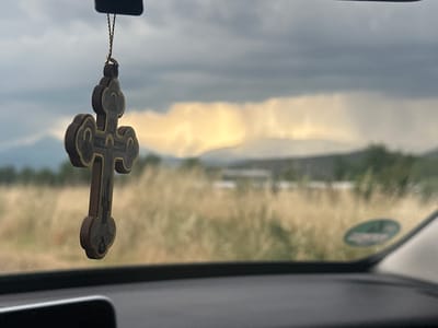 The image presents a view from inside a car, looking out through the front windshield at an expansive landscape. In the immediate foreground and centered, a wooden Orthodox cross hangs from what appears to be the rearview mirror. The cross is intricately carved with religious figures and text, including visible Greek letters "IC XC" (Jesus Christ) and "NI KA" (conquers).
Beyond the car's dashboard, which occupies the lower part of the frame, lies a field of dry, light-colored grass or crops, stretching towards distant mountains. The sky is largely overcast with dark, heavy clouds, but a striking band of golden light illuminates the horizon, suggesting sunlight breaking through a specific area of clouds, possibly indicating localized rain or a dramatic change in weather. This creates a powerful contrast between the dark clouds and the glowing distant landscape.
The overall light suggests it is daytime, with the weather appearing cloudy and potentially stormy in the distance. A blurry green circular sticker is visible on the right side of the windshield. The scene depicts a journey through a rural or semi-rural area, consistent with the location context of Bogdanci, North Macedonia, with the cross signifying faith or protection during travel.