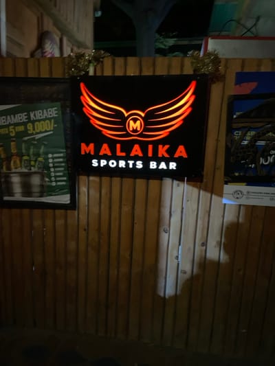 The image shows a sign for a sports bar called "Malaika Sports Bar" located in Dodoma, Tanzania (latitude: -6.165, longitude: 35.745). The sign is a black metal rectangle with red neon lettering and an illuminated logo of wings and the letter "M" inside. The sign is mounted on a wooden wall, which is also decorated with two other posters on the left and right. The sign is positioned in a dimly lit environment. There are no people present in the image. The wooden wall is made of vertical planks. The lighting source is coming from the sign's neon lettering and logo. The sign's shadow can be seen on the wall behind it.