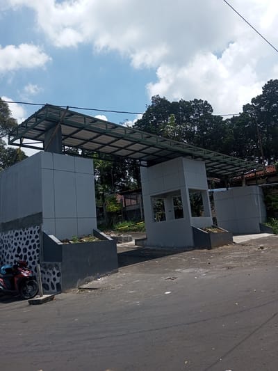 The image depicts a newly constructed or renovated entrance structure, possibly for a residential area or public space. The structure consists of gray concrete pillars and walls, connected by a metal roof frame. The pillars have small planters at their bases. One of the pillars has an open section with windows, suggesting it could be a guardhouse or checkpoint.
A red motorcycle is parked near one of the pillars. The road surface in front of the structure is asphalt and appears to be well-used. The background features trees, vegetation, and possibly some residential buildings.
The location is unknown and there is no basis to assume it is Kioumaknao 2, Laos.