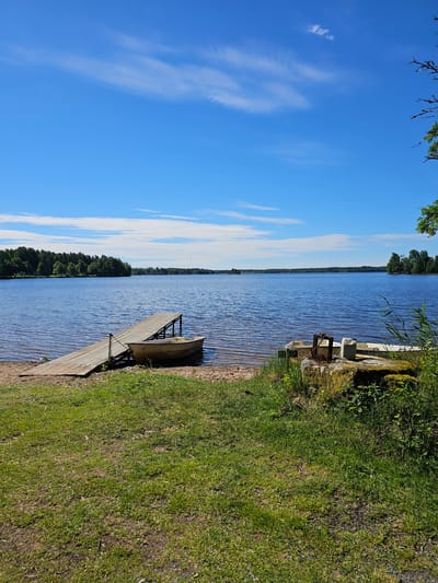 The image depicts a tranquil lake scene under a bright blue sky with scattered white clouds. A wooden pier extends from a sandy shore into the calm, blue water. A small, light-colored boat is moored to the pier. The shore is lined with green grass and some vegetation. In the background, a dense line of trees borders the opposite side of the lake, suggesting a natural, possibly rural or park-like setting. On the right side of the foreground, a large rock with some moss and plants is visible, with a white plastic container and metal object resting on it. This scene is likely located near Nässjö, Sweden, given the context provided.