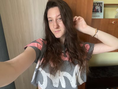 In the city of Popintsi, Bulgaria, a young woman is taking a selfie in a room. She has long, dark brown hair and is wearing a gray t-shirt with pink and white abstract designs. She is smiling slightly and her right hand is touching her hair. In the background, there is a wooden wardrobe with a shelf containing a framed photo of several people and some decorative items. A television is visible below the shelf. The room appears to be well-lit, with a light-colored wall and a darker surface on the right.