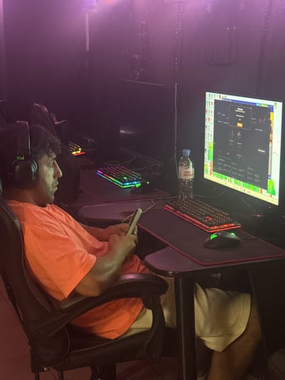 A man wearing headphones and an orange t-shirt sits in a gaming chair, holding a mobile phone in his right hand. He is facing a computer monitor displaying a game interface. A keyboard with RGB lighting is visible in front of the monitor, along with a gaming mouse. A bottle of "Earn Spring" water stands next to the monitor. The environment appears to be a dimly lit room, possibly an internet cafe, with purple lighting. The image is shot from a medium side angle, capturing the man's profile. The scene is located in Beryozovka, Russia.